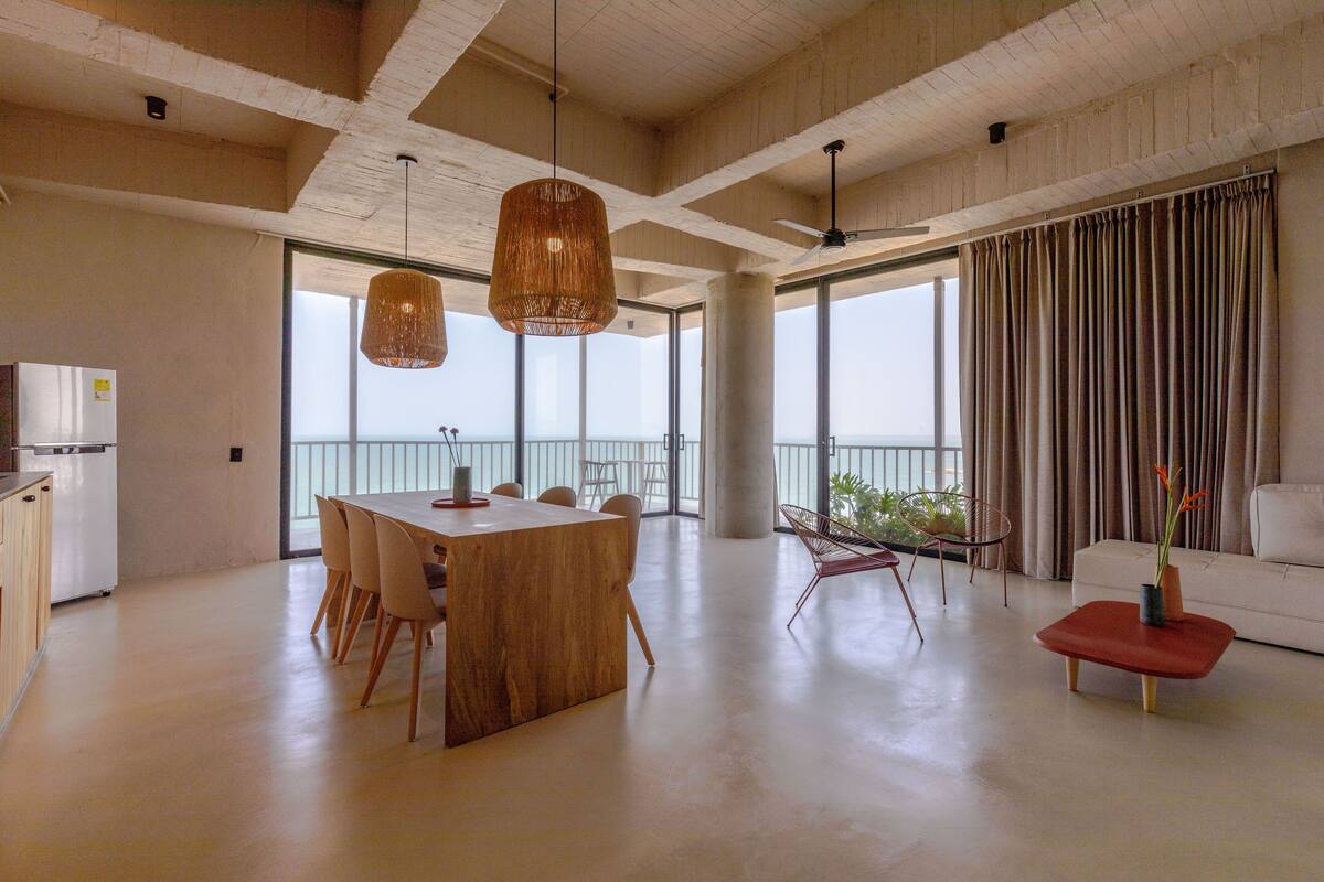 A spacious living area with a wooden dining table, chairs, a red ottoman, a white sofa, and a view of the ocean through large windows.