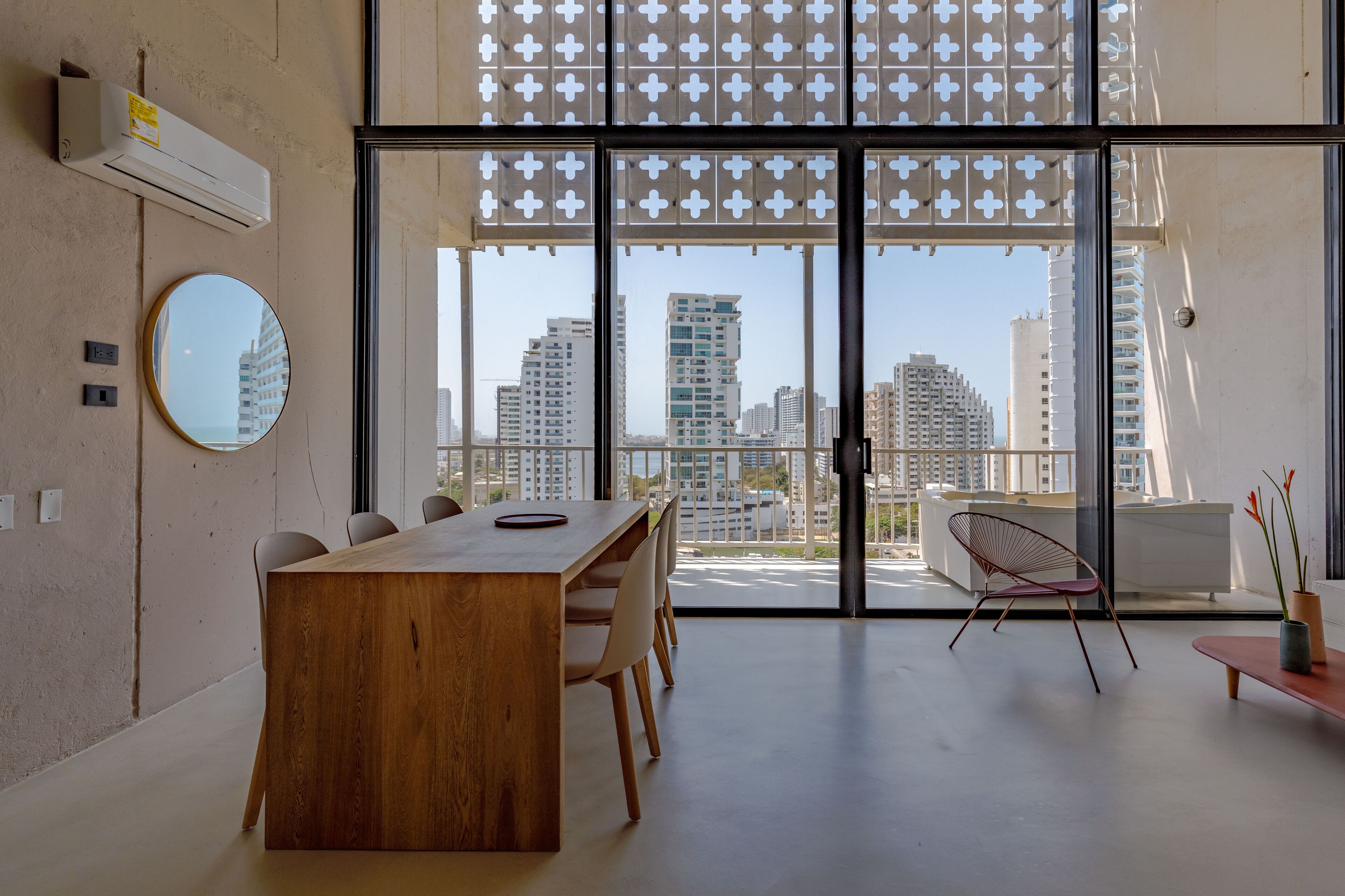 A modern dining area with a wooden table and chairs, a round mirror, and a view of tall buildings through large glass doors.