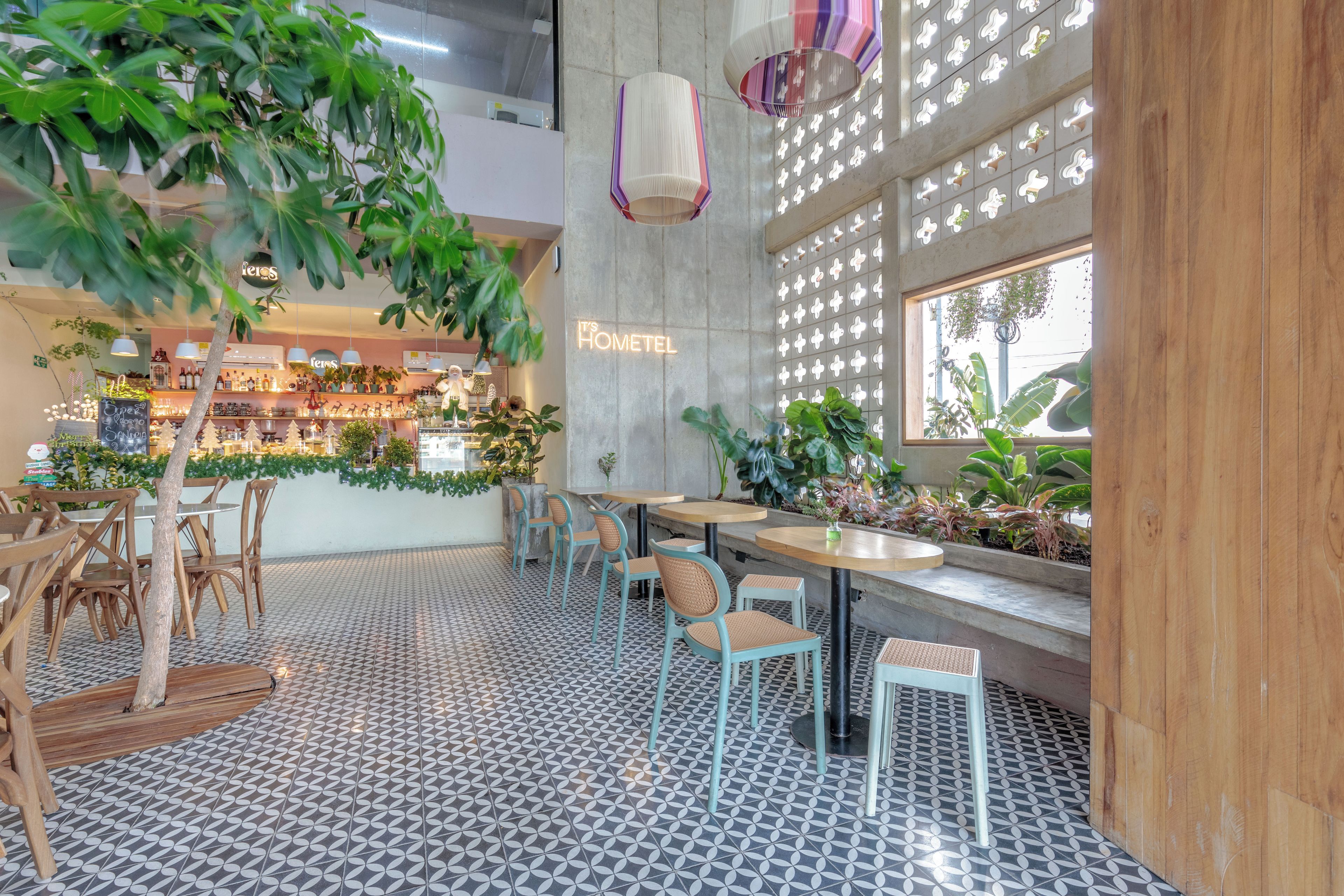 A cafe interior with a patterned floor, wooden tables and chairs, hanging plants, and a wall with a sign that reads 'IT'S HOMETEL'.