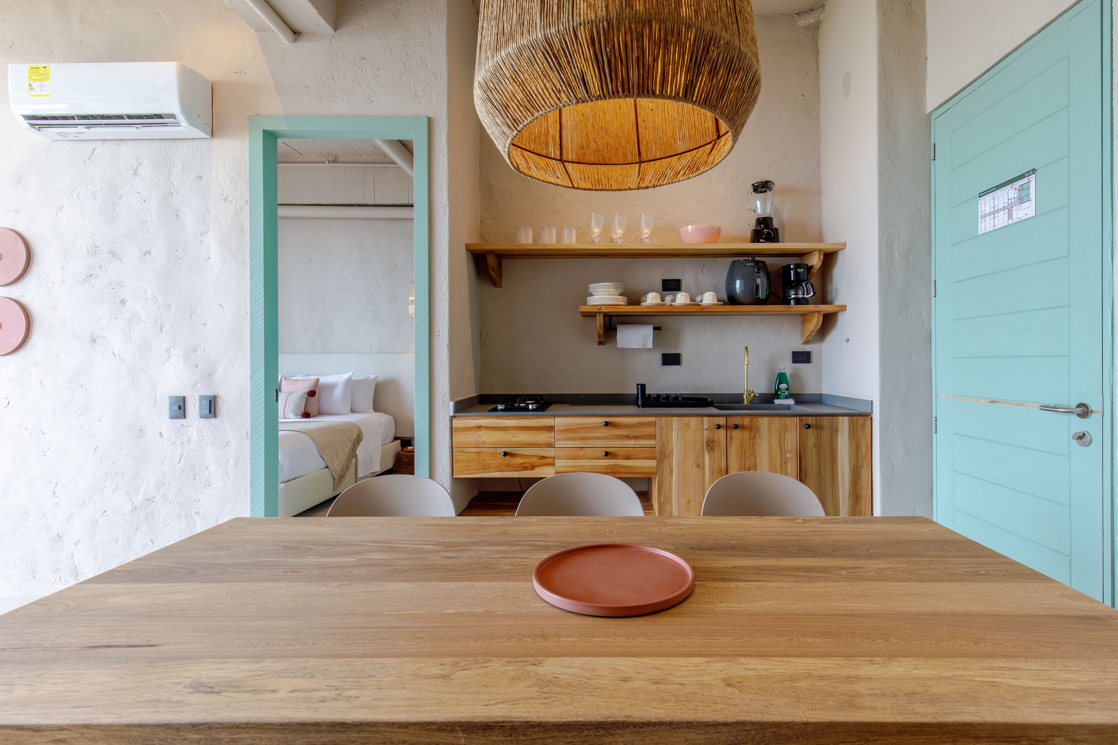 A modern kitchen with wooden cabinets, a dining table with a red plate, and a hanging wicker light fixture.