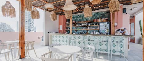 Uma área de estar ao ar livre com cadeiras e mesas brancas, um bar com balcão de padrão verde e luminárias trançadas penduradas.