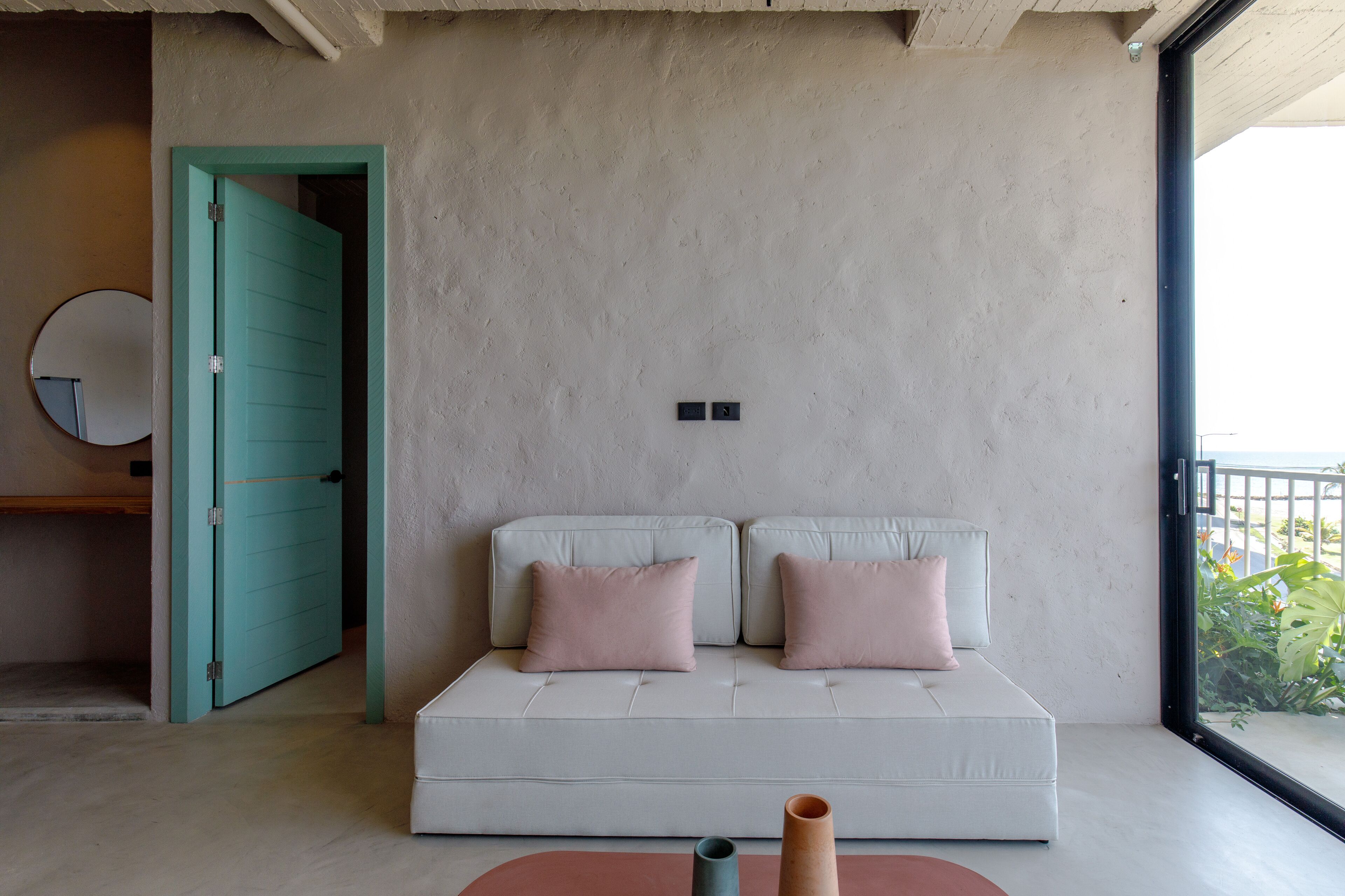A modern living room with a white sofa, two pink cushions, a round mirror, and a small table with two vases.