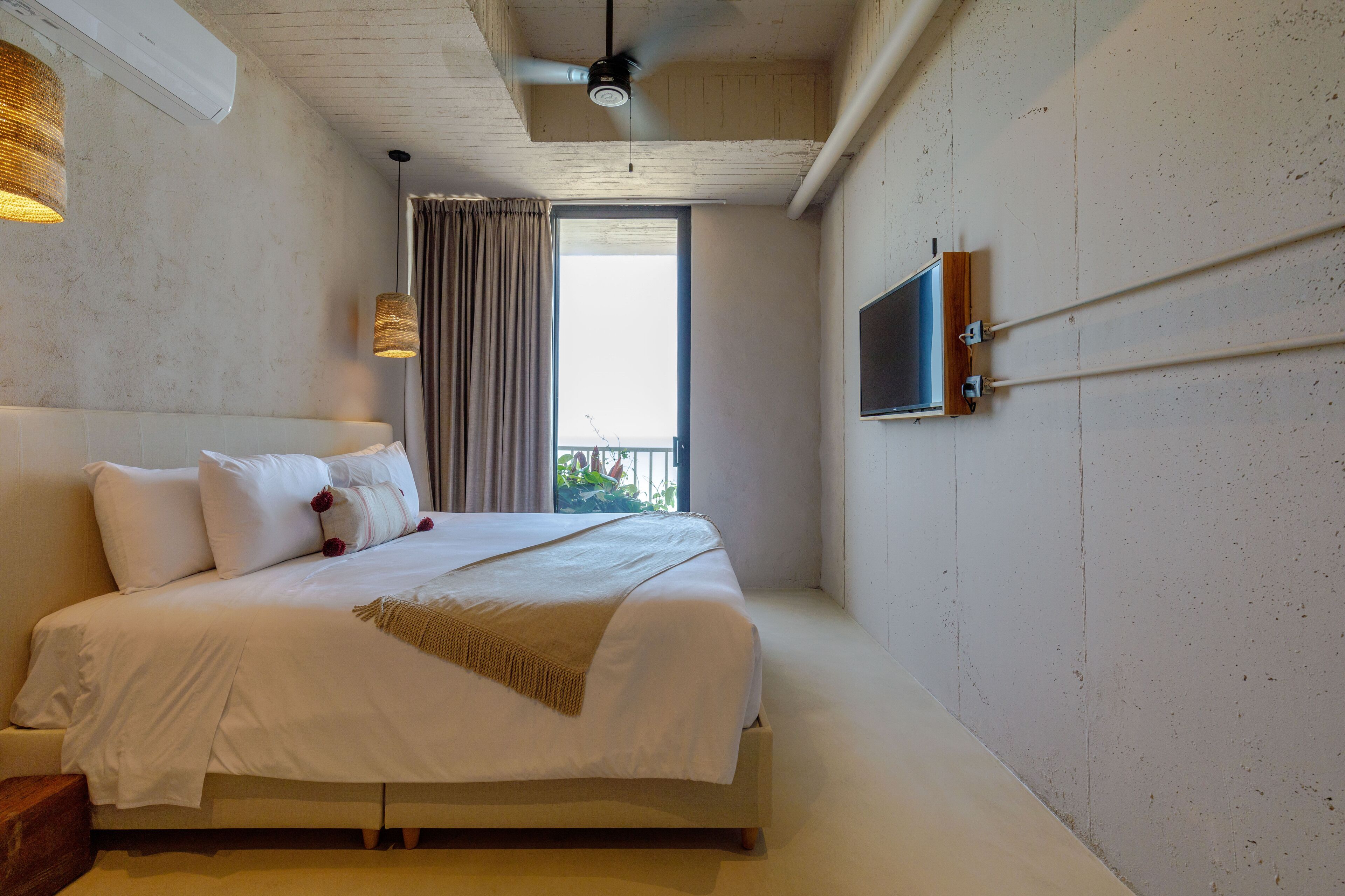 A bedroom with a large bed, a ceiling fan, a TV mounted on the wall, and a window with curtains.