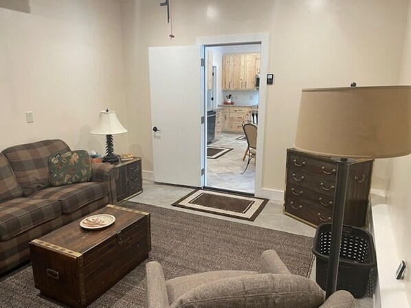 A living room with a sofa, a coffee table, a lamp, and a chair. There is a door leading to a kitchen area. - Trails End Lodge -comfort, convenience & location -10% discount for 7 night stay (Trout Lake)