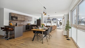 A modern kitchen with a central island, a dining area with a wooden table and chairs, and a view of the outside through large windows. - GebirgsResort Winterberg (Winterberg)