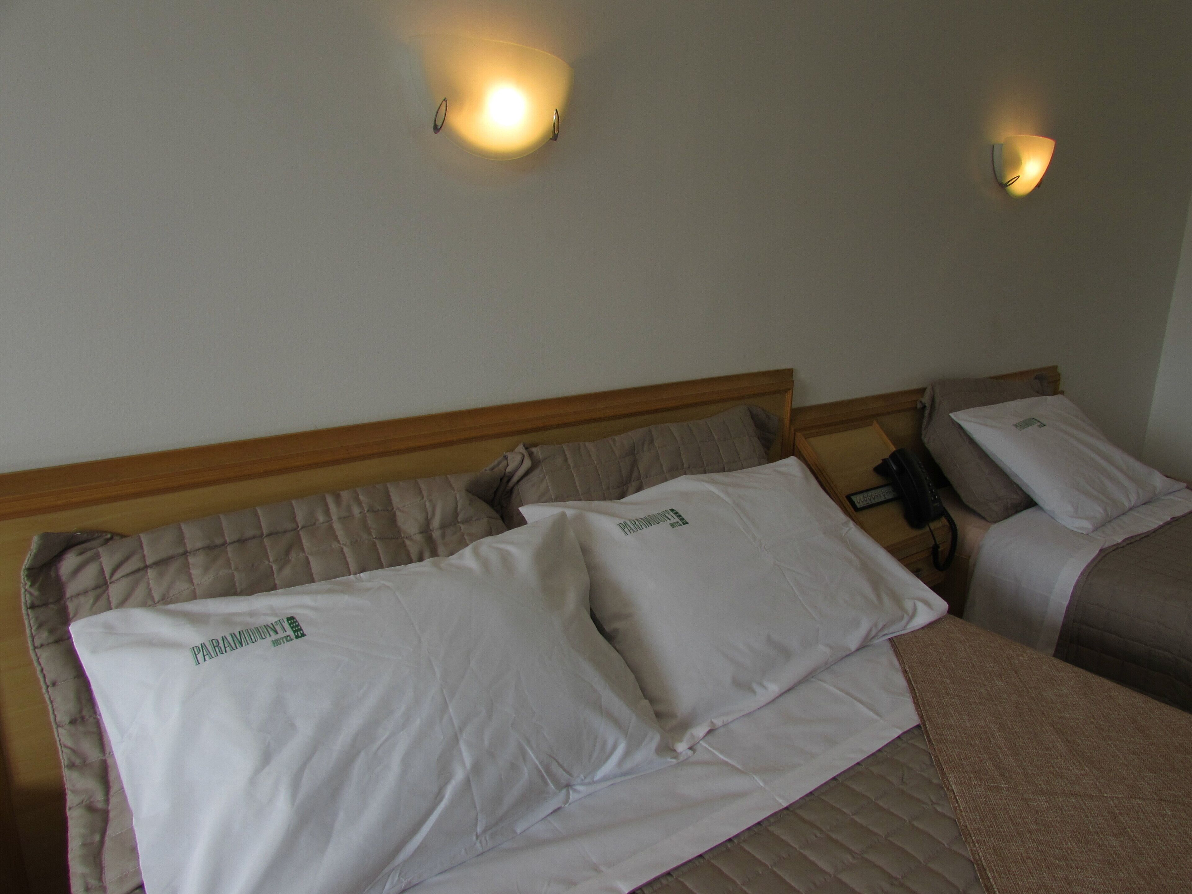 A hotel room with two beds, each with a pillow labeled "Paramount Hotel." There are wall-mounted light fixtures above the beds.