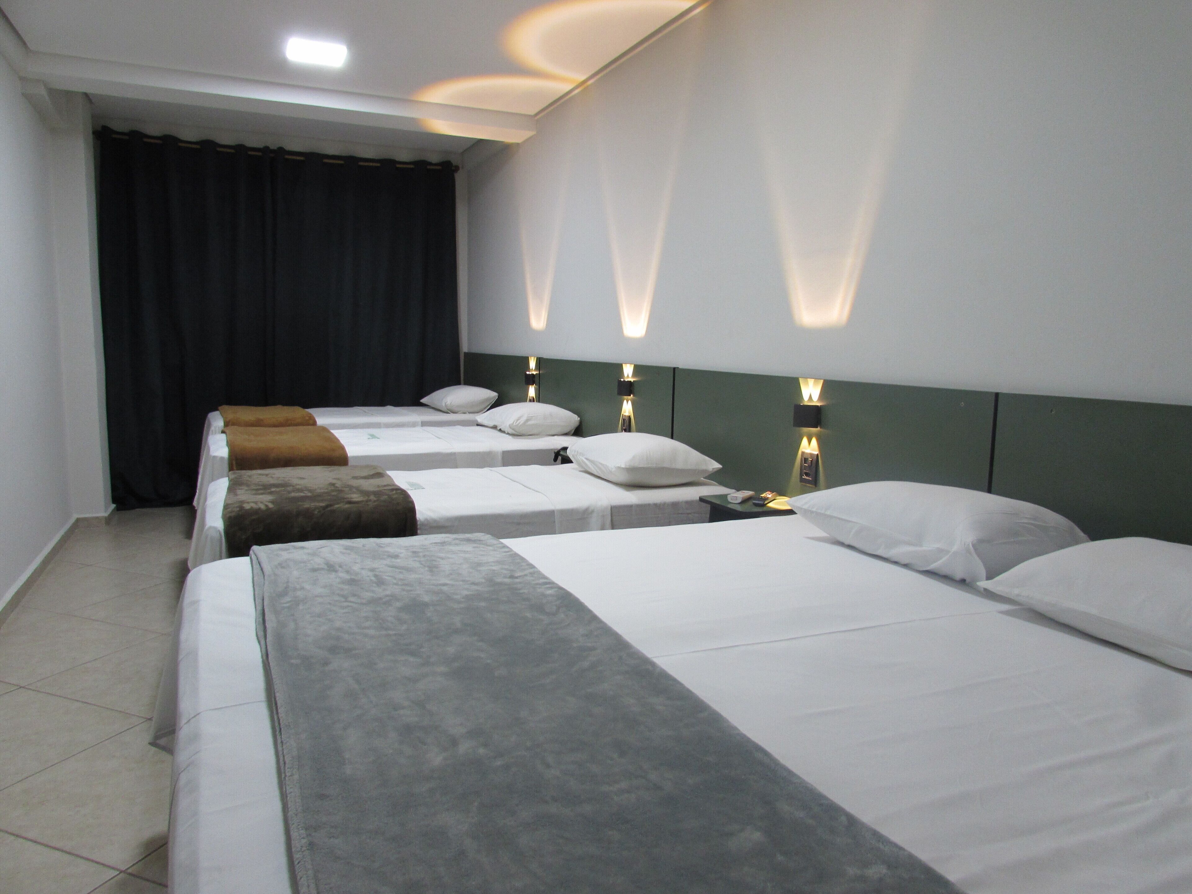 A hotel room with four single beds, each with a pillow and a blanket. There are wall-mounted lights above each bed.