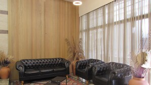 Una habitación con un sofá de cuero negro, un sillón a juego y una pequeña mesa. Hay una alfombra estampada en el suelo y un ventanal con cortinas translúcidas.
