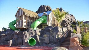Ein Wasserpark mit einer Rutsche, einem Gebäude mit Reetdach und einem felsigen Hintergrund.