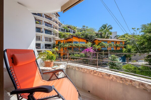 A balcony with a lounge chair and a potted plant, overlooking a building with balconies and a stone wall.