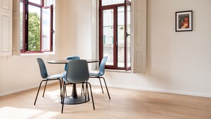 Un comedor con una mesa redonda y cuatro sillas, ventanas amplias con contraventanas abiertas y un cuadro en la pared.