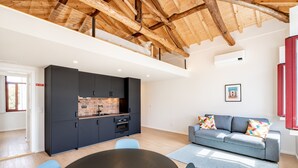 Una cocina moderna con armarios oscuros, salpicadero de ladrillo y techo de madera. En la zona de estar, hay un sofá gris con cojines de colores.