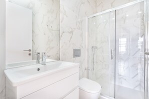 Un baño moderno con lavamanos, espejo y retrete blancos, además de una cabina de ducha de vidrio.