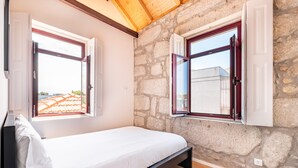 Un dormitorio con cama, paredes de piedra y dos ventanas con contraventanas abiertas.