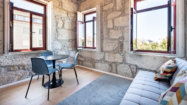 Una habitación con paredes de piedra, techo de madera, una mesa redonda con sillas, un sofá con un cojín de colores y ventanas grandes con marcos rojos.