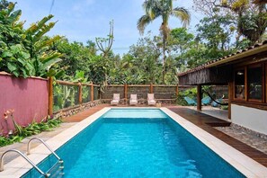 Piscina rectangular con cubierta de madera y sillas de descanso, rodeada de vegetación exuberante y una cerca de madera.