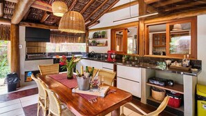 A rustic kitchen with wooden beams, a dining area with wicker chairs, and a table set with wine glasses and bottles. - Praia de Itamambuca - Bali House (Ubatuba)