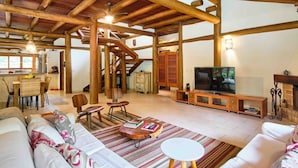 A spacious living area with a wooden ceiling, a television, a fireplace, and a dining area with a kitchen in the background. - Praia de Itamambuca - Bali House (Ubatuba)