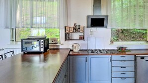 A modern kitchen with a built-in oven, a kettle, and a coffee machine. There are two windows with sheer curtains, and a small shelf with coffee bags and bottles. - 4 bedroom beautiful home in Suleczyno (Suleczyno)