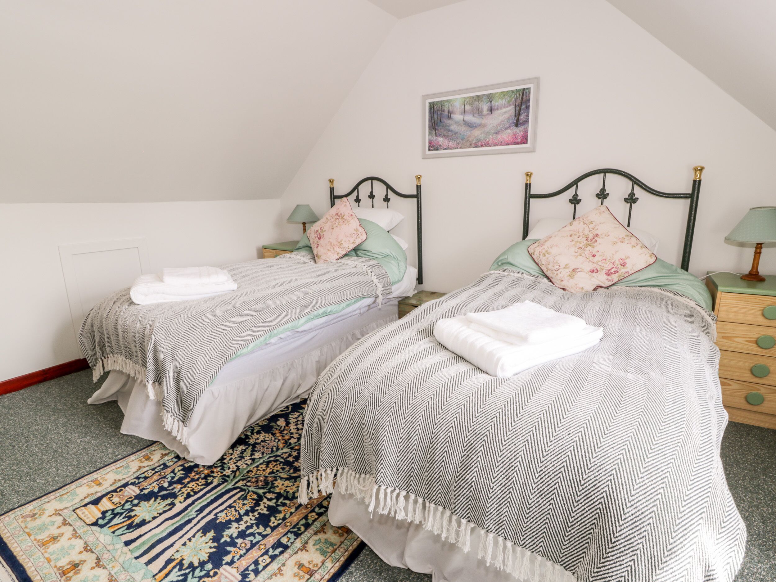 A bedroom with two beds, a patterned rug, a painting, and a nightstand with a lamp.