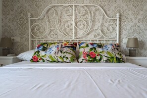 A neatly made bed with white bedding and floral pillows, a white metal headboard, and a patterned wallpaper background.
