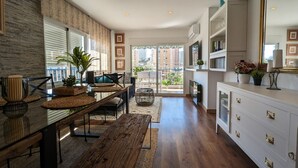 A spacious living area with a wooden dining table, comfortable seating, a large window with a view of buildings, and a white cabinet with drawers.
