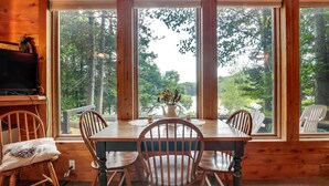 A wooden cabin with a dining table, four chairs, and a large window offering a view of trees. - Lakefront Ogema Vacation Rental Cabin w/ Fire Pit! (Ogema)