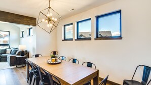 Uma área de refeições com mesa de madeira, cadeiras pretas e uma luminária pendente geométrica. Há três janelas na parede, e é visível uma casa através delas.