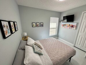 Quarto com cama, almofadas, mesa de cabeceira com abajur, quadros nas paredes, televisão e pôster motivacional.