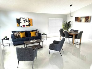 A modern living room with a sofa, coffee table, and chairs, a dining area with a table and chairs, and artwork on the walls.
