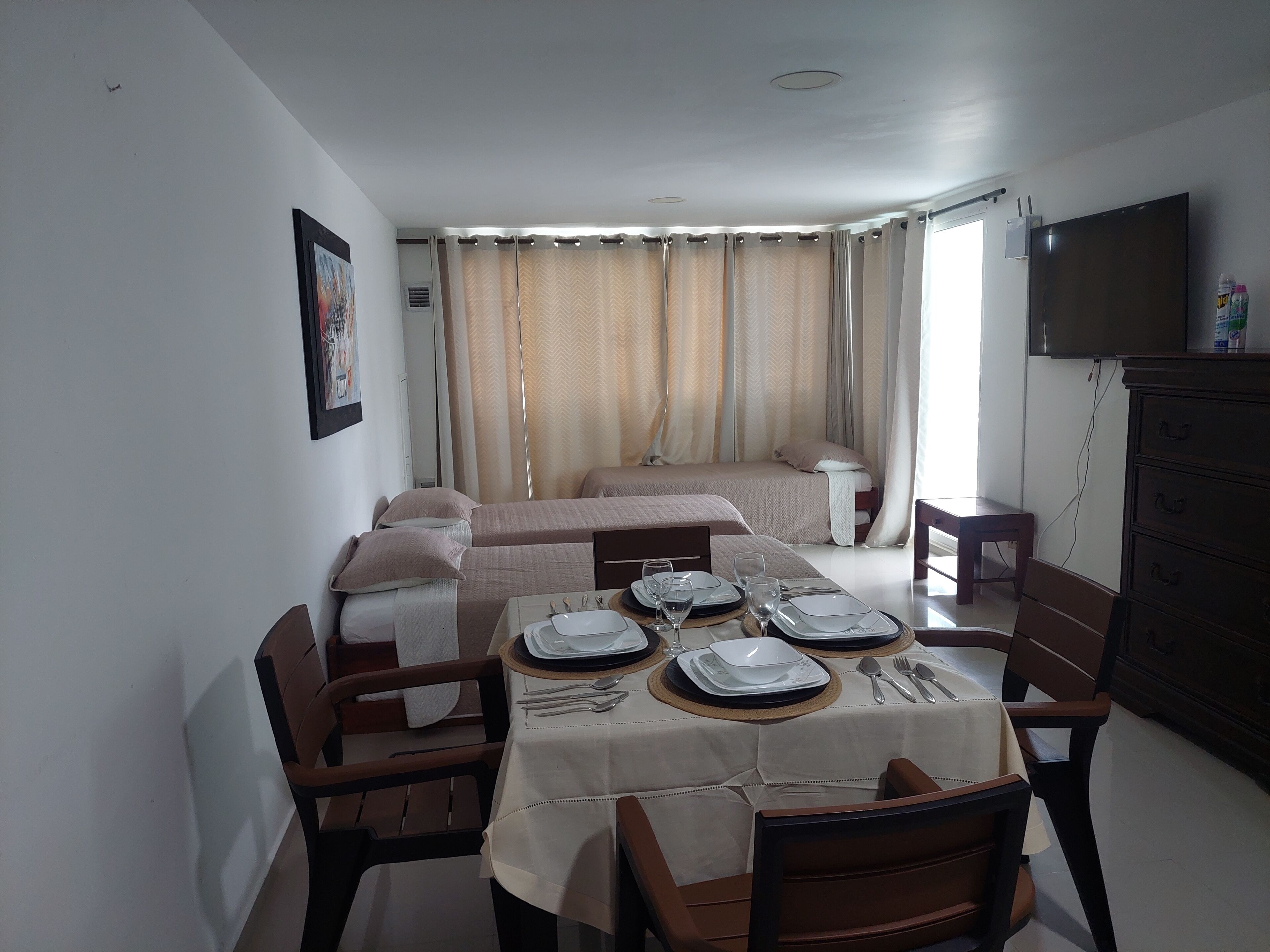 Une chambre d’hôtel avec un coin repas aménagé pour deux personnes, un lit doté de rideaux, un téléviseur fixé au mur et une petite table avec une bouteille.