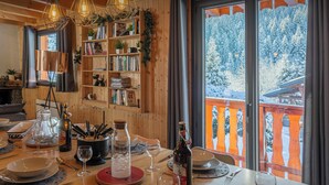 Un coin repas chaleureux avec une table en bois dressée pour le repas, une bibliothèque remplie de livres et une vue sur un paysage enneigé par de grandes fenêtres.