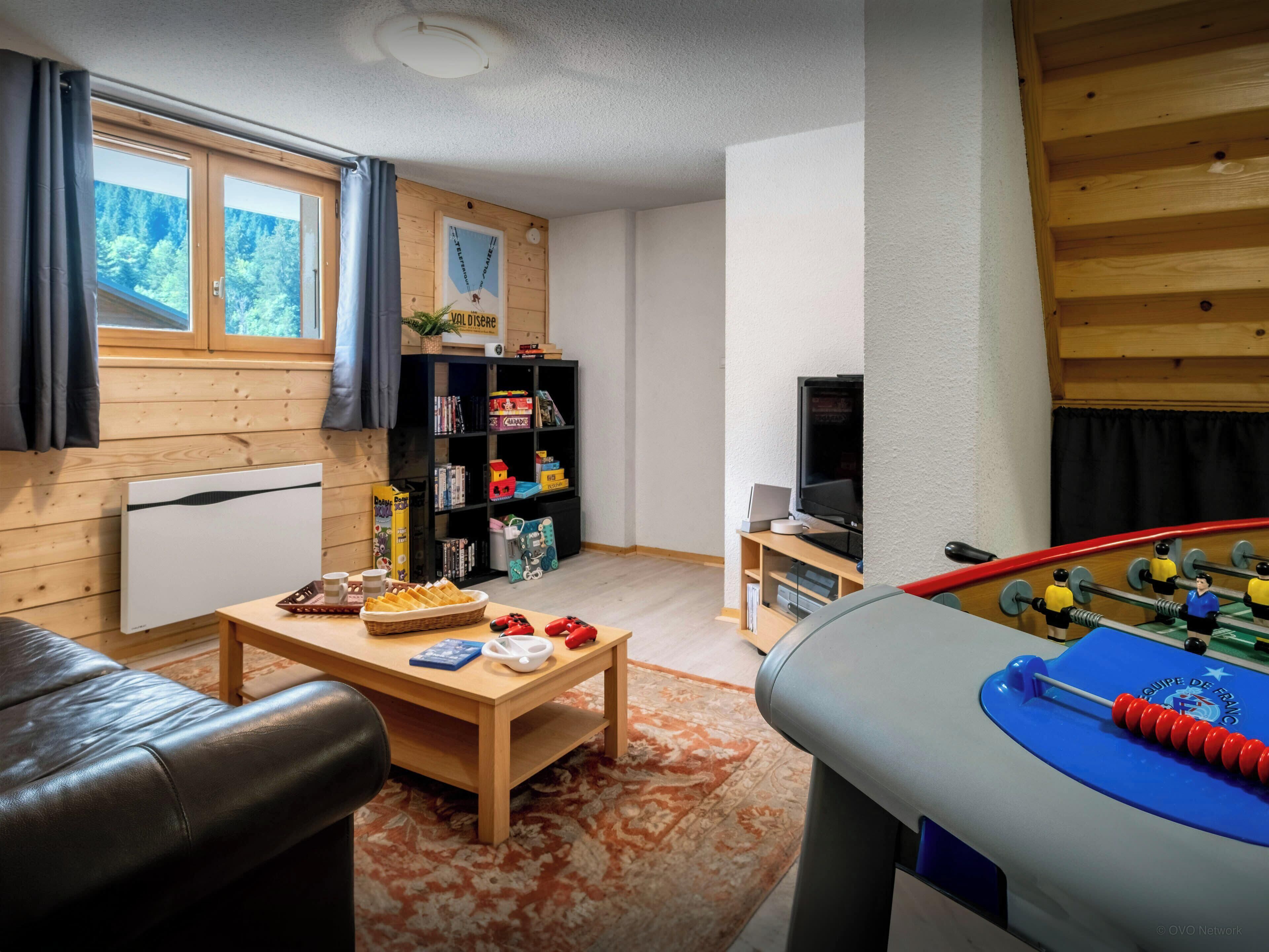 A cozy living room with a foosball table, a television, a bookshelf, and a window with curtains.