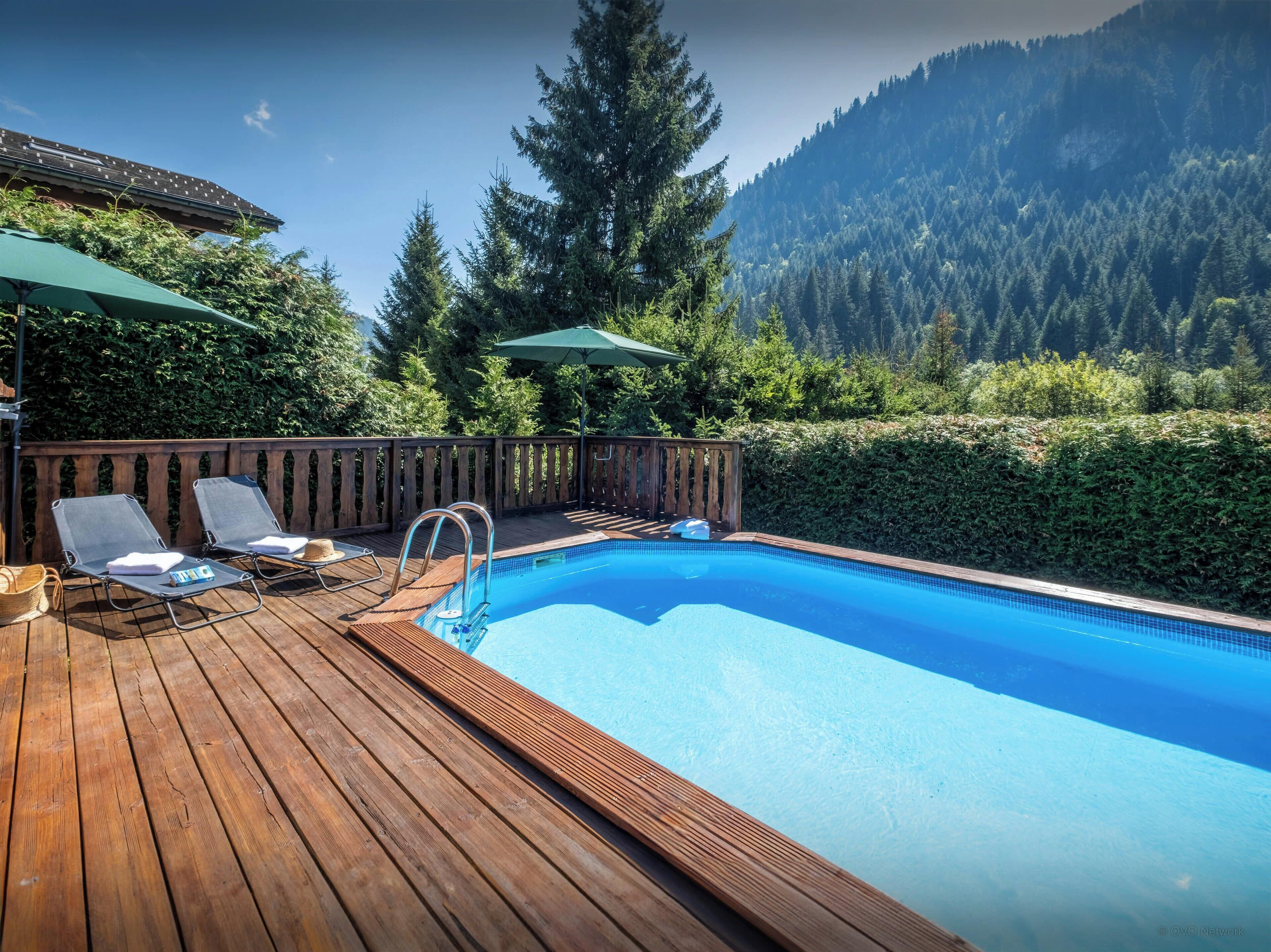 Une terrasse en bois avec une piscine, deux chaises longues et une petite table sur laquelle se trouvent un chapeau et une serviette.