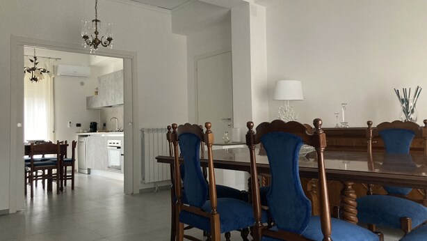 Un comedor con una mesa de madera y sillas tapizadas en azul.  Hay una araña de luz sobre la mesa, y se ve una cocina al fondo.