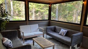 A covered outdoor seating area with two sofas and a wooden table, surrounded by large windows offering a forest view.