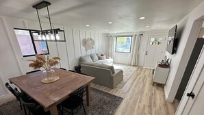 A modern living room with a wooden dining table, a sofa, and a TV mounted on the wall. - Heart of Richfield, ATVs & Mountain Biking, Spacious Home, Sporting Events (Richfield)