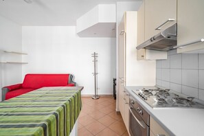 A compact room with a kitchenette, a bed with a red headboard, and a standing heater.