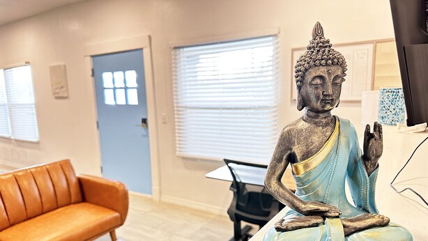 Eine Statue einer sitzenden Figur in einem Raum mit einem braunen Ledersofa, einem Holztisch und einer Tür mit einem Fenster.