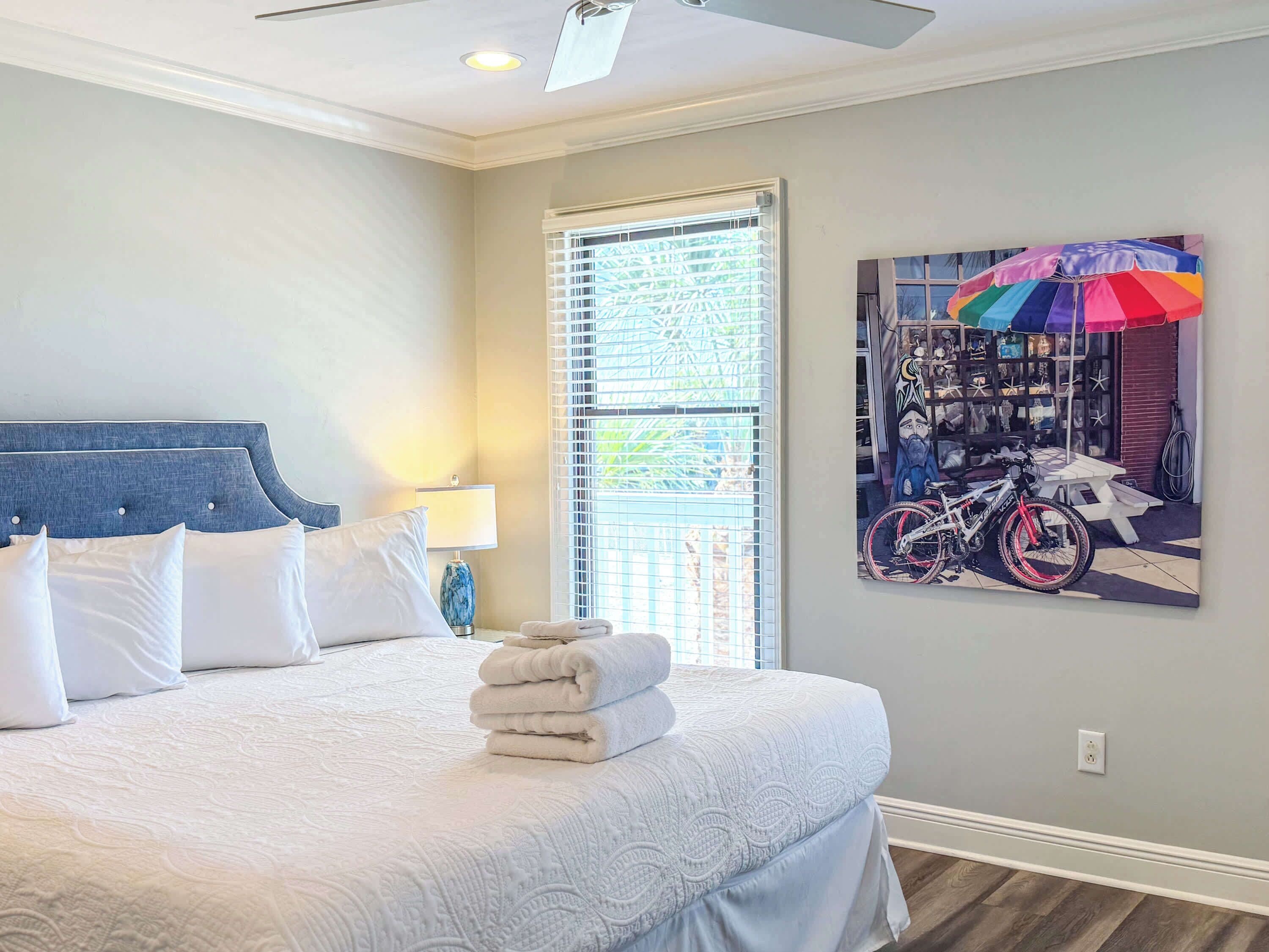 A bedroom with a bed, a window with blinds, a ceiling fan, and a wall-mounted artwork.