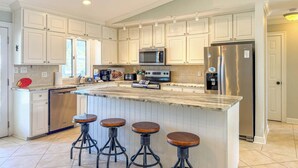 A kitchen with white cabinets, a stainless steel refrigerator, a microwave, and a coffee maker. There are four stools at the island.