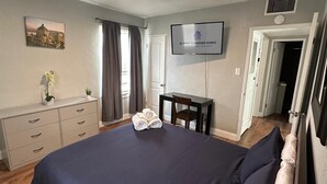Een verzorgde slaapkamer met een bed, een bureau, een stoel, een televisie en een raam met gordijnen.