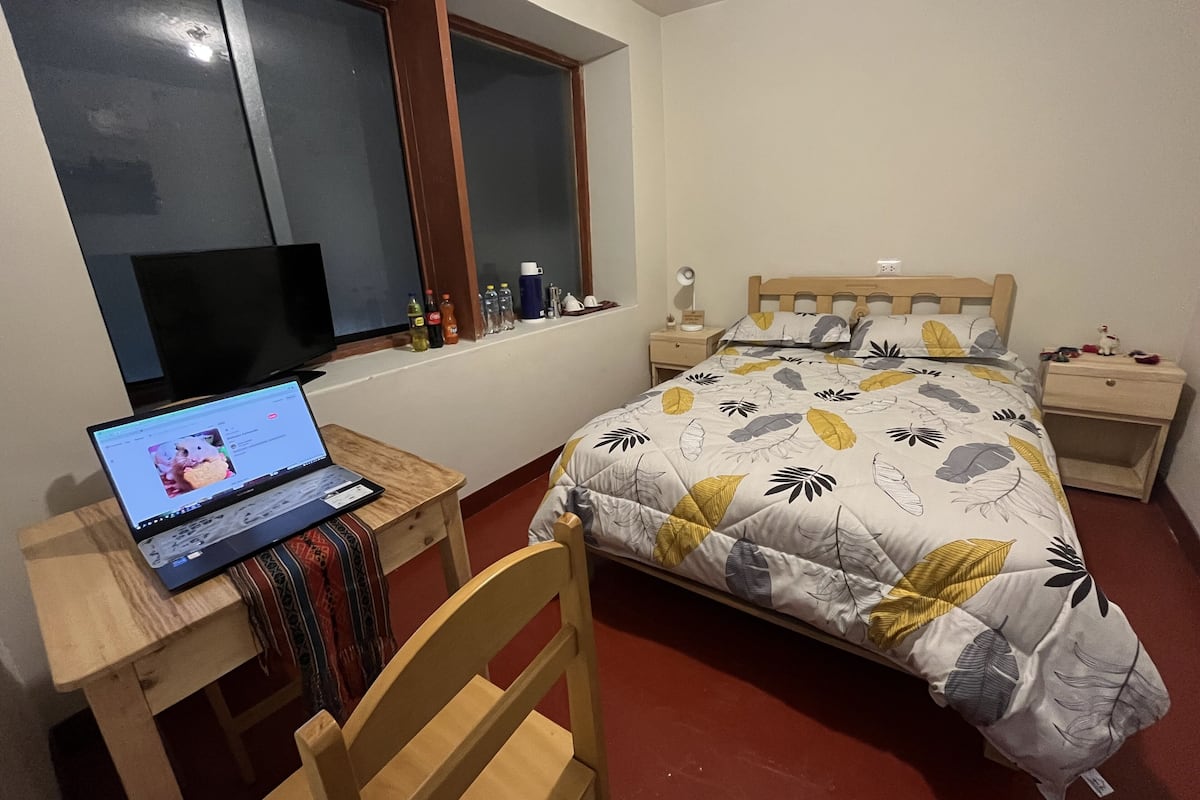 Ein Schlafzimmer mit einem Bett, einem Schreibtisch mit Laptop, einem Stuhl, einem Nachttisch und einem Fenster mit Jalousien.