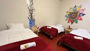 A hotel room with three single beds, each with a red blanket and a pillow. There is a wooden nightstand with a bottle and a small item on it. A colorful wall decoration is visible on the right wall. - The Sleepy Mouse (Cusco)