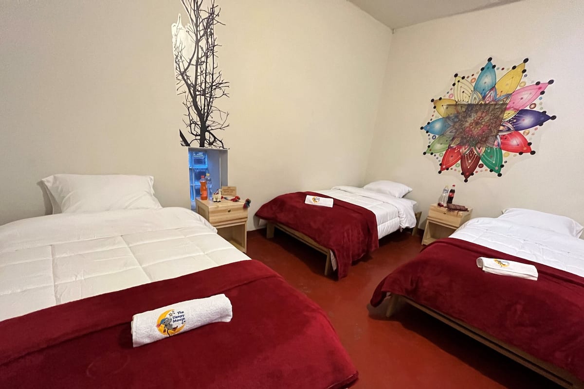 Ein Hotelzimmer mit drei Einzelbetten, jedes mit einer roten Decke und einem Kopfkissen. Ein hölzerner Nachttisch zeigt eine Flasche und ein kleines Objekt. An der rechten Wand ist eine farbenfrohe Wanddekoration zu sehen.