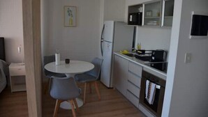 Uma cozinha compacta com geladeira, micro-ondas e forno. Há uma mesa de jantar com cadeiras e um pequeno armário acima do balcão.