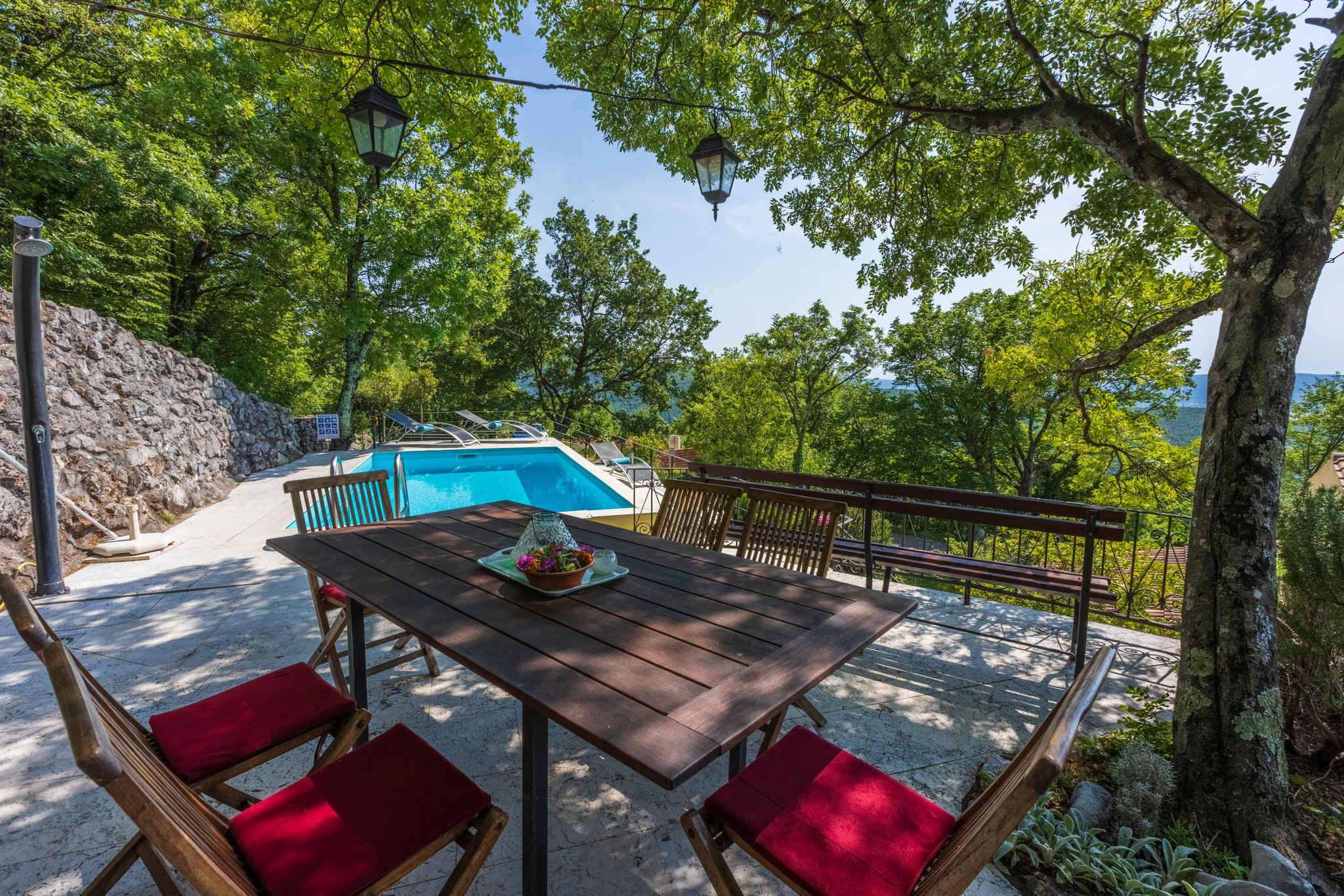 An outdoor dining area with a wooden table and red cushioned chairs, overlooking a swimming pool and a rocky hillside.