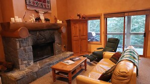 A cozy living room with a stone fireplace, leather sofa, and a large window offering a view of trees. - Ski Bike in out 4 bdrm, 4 bth home w pool table & media room, plus 2 sofa beds (Whistler)