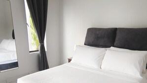 A neatly made bed with white linens and two dark pillows, a bedside table with a lamp, and a window with curtains. - Paddy Villea Inn (Bukit Mertajam)