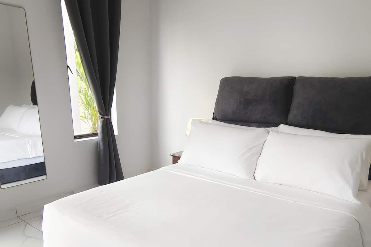 Un lit bien fait, avec du linge de lit blanc et deux oreillers foncés, une table de chevet avec une lampe et une fenêtre avec des rideaux.